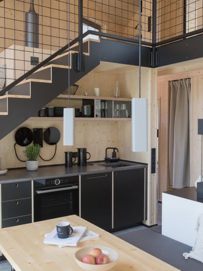 Eine moderne und minimalistische Küche mit schwarzen Schränken und Holzdetails. Im Vordergrund steht ein Holzstisch mit einer Schale Äpfeln und einer Tasse.