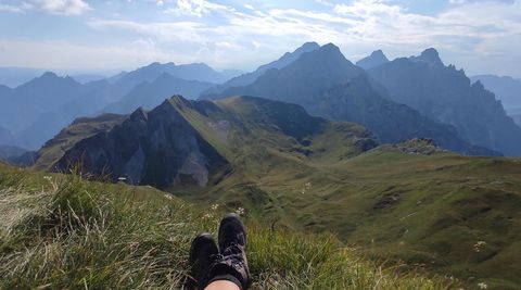 Eine entspannende Aussicht auf hochgelegene Berge und sanfte Hügel. Im Vordergrund befinden sich Füße in Wanderstiefeln auf grünem Gras.