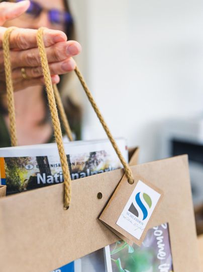 Une main tient un sac en papier avec une étiquette et plusieurs brochures. Le sac a un design simple et élégant.