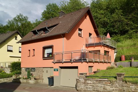 Ein modernes, orangefarbenes Haus mit einem Balkon und einer Garage. Es steht in einer grünen Umgebung zwischen anderen Wohnhäusern.