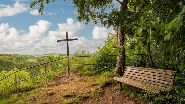 Eine Holzbank steht an einem Aussichtspunkt mit einem Kreuz und Blick auf die grüne Landschaft. Die Wolken am blauen Himmel verleihen der Szene ein friedliches Ambiente.