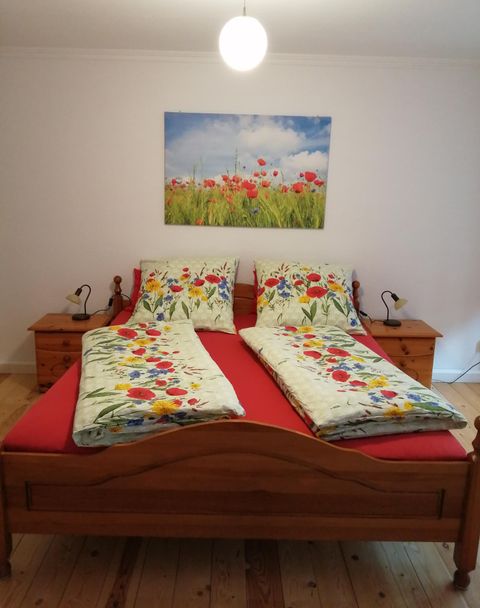 Ein gemütliches Schlafzimmer mit einem Holzbett und bunten Bettwäsche. An der Wand hängt ein Bild von blühenden Blumenfeldern.