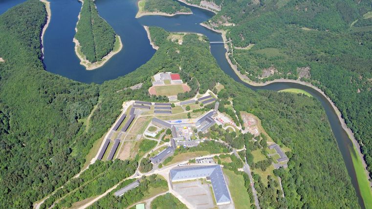 Eine luftige Aufnahme eines Flusses, der sich durch üppiges Grün schlängelt. Im Vordergrund sind Gebäude und ein weitläufiges Gelände zu sehen.
