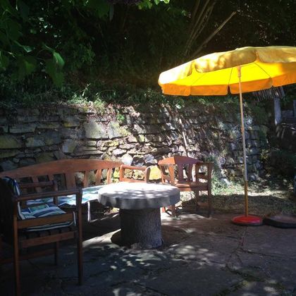 Ein gemütlicher Sitzbereich im Freien mit Holzmöbeln und einem Sonnenschirm. Im Hintergrund sind Steinmauern und Pflanzen zu sehen.