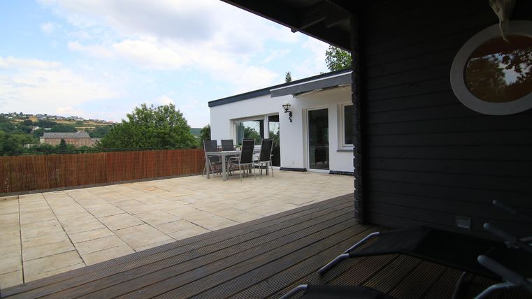 Eine großzügige Terrasse mit einem modernen Haus im Hintergrund. Der Blick geht auf eine grüne Landschaft unter einem klaren blauen Himmel.