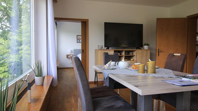 Ein moderner Essbereich mit einem großen Tisch und gemütlichen Stühlen. Im Hintergrund ist ein Fernseher und eine helle Fensterfront mit Blick ins Grüne.