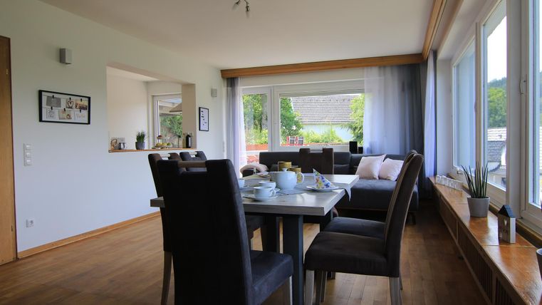 Ein modernes Wohnzimmer mit einem großen Tisch und bequemen Stühlen. Durch die großen Fenster fällt viel Licht und bietet einen Blick ins Grüne.