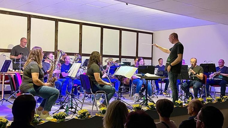 Ein Orchester spielt auf einer Bühne, während der Dirigent die Musiker anleitet. Verschiedene Instrumente sind zu sehen, und das Publikum sitzt im Vordergrund.