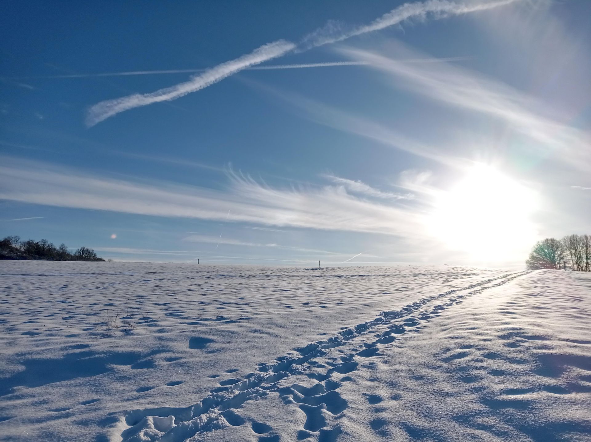 Eine verschneite Landschaft unter einem klaren blauen Himmel. Die Sonnenstrahlen scheinen hell und warme Fußspuren führen durch den Schnee.