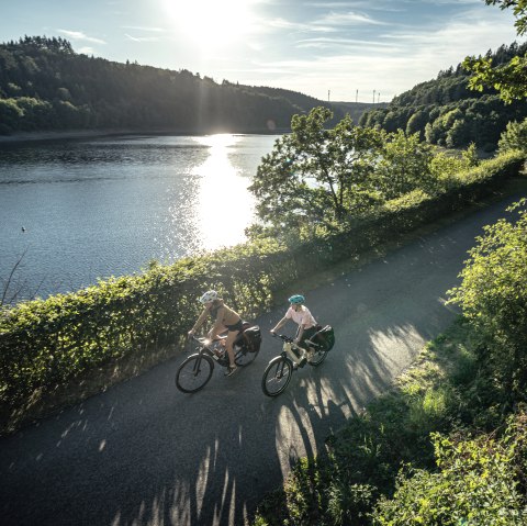 Zwei Radfahrer fahren bei Sonnenschein entlang eines Sees, umgeben von grüner Natur. Die Sonne spiegelt sich im Wasser., © Eifel Tourismus GmbH, Dennis Stratmann