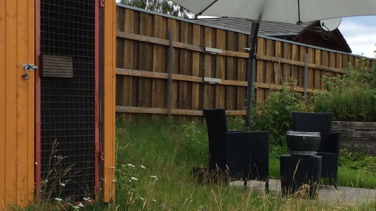 Ein gemütlicher Außenbereich mit zwei Stühlen und einem Tisch unter einem Sonnenschirm. Im Hintergrund steht eine orangefarbene Hütte und ein Holzzaun.