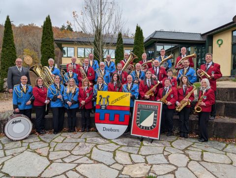 Eine große Blaskapelle steht im Freien und posiert für ein Gruppenfoto. Die Musiker tragen farbige Uniformen und halten ihre Instrumente.