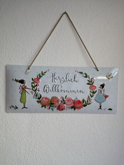 Ein dekoratives Schild mit der Aufschrift „Herzlich Willkommen“. Es zeigt zwei Mädchen und eine florale Umrahmung mit Rosen.