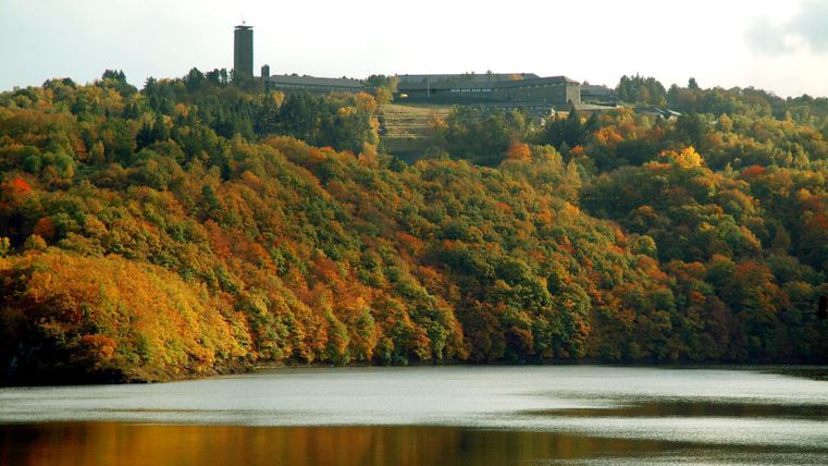 Eine malerische Landschaft mit buntem Herbstwald und einem ruhigen See. Im Hintergrund ist ein Gebäude auf einem Hügel zu sehen.