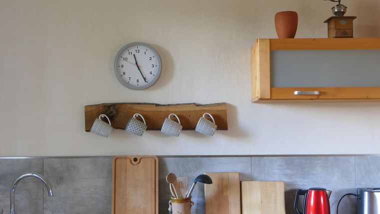 Eine gemütliche Küche mit einem hölzernen Regal, auf dem vier Tassen hängen. An der Wand sind eine Uhr und einige Kochutensilien sichtbar.