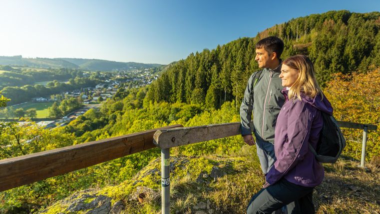 Ein Paar steht an einem Aussichtspunkt und blickt auf eine grüne Landschaft. Im Hintergrund sind Bäume und kleine Häuser zu sehen.