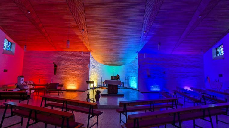 Eine moderne Kirche mit bunten Lichtern in Rot, Blau und Violett. Bänke sind in einem offenen Raum angeordnet, der eine ruhige Atmosphäre ausstrahlt.