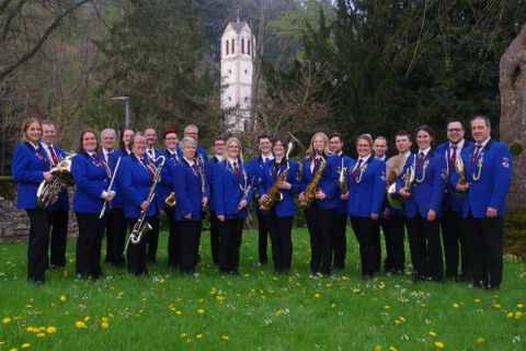 Eine Gruppe von Musikern steht im Freien in eleganten blauen Jacken. Im Hintergrund ist ein Gebäude zu sehen, umgeben von einer grünen Landschaft.