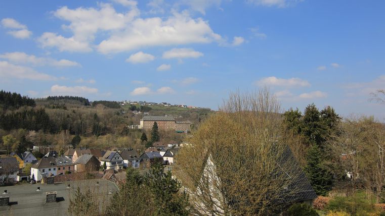 Eine ländliche Landschaft mit mehreren Häusern und sanften Hügeln im Hintergrund. Der Himmel ist blau mit wenigen Wolken.