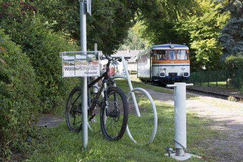 Ein Fahrrad steht an einem Parkplatz mit einem Schild für den Radweg. Im Hintergrund fährt ein Zug vorbei.
