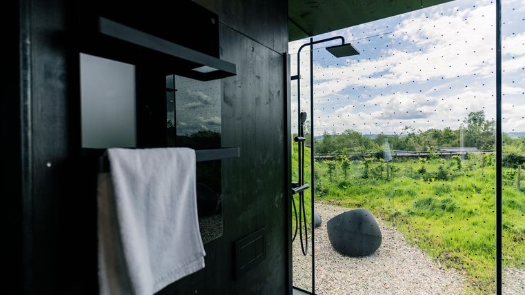 Ein modernes Badezimmer mit dunklen Wänden und einem Handtuch. Im Hintergrund sieht man eine große Glaswand, die einen Blick auf die grüne Landschaft ermöglicht.