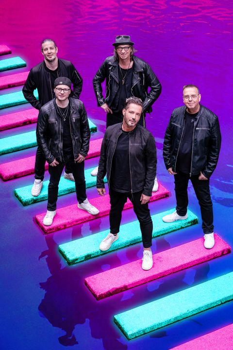 Eine Band steht auf bunten Plattformen über lila Wasser. Alle Mitglieder tragen schwarze Kleidung und sehen stylisch aus.