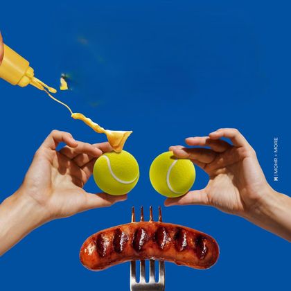 Zwei Hände halten Tennisbälle und eine Wurst, während Senf darüber verteilt wird. Der Hintergrund ist leuchtend blau.