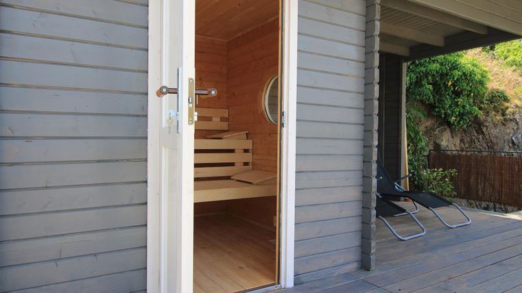 Eine moderne Sauna mit Holzinterieur und offenem Eingang. Neben der Sauna stehen zwei Liegen auf einer Terrasse.