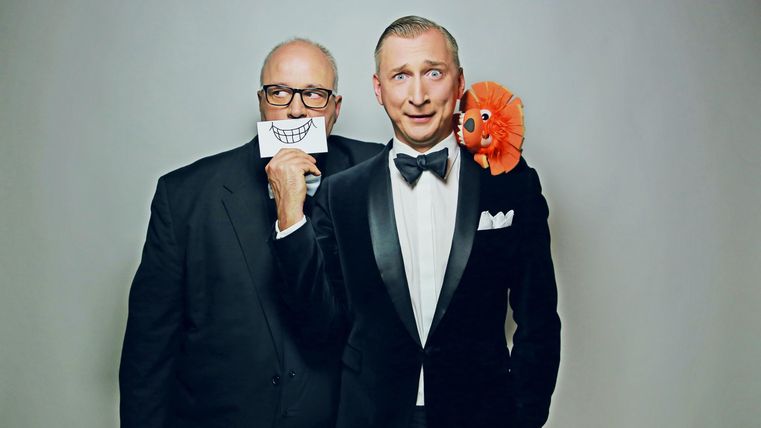 Zwei Männer in formeller Kleidung stehen zusammen. Der Mann vorne hält ein großes Lächeln-Schild und hat einen orangefarbenen Plüschkopf auf seiner Schulter.
