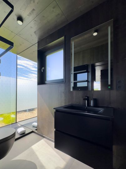 Ein modernes Bad mit schwarzen Möbeln und großen Fenstern, die viel Tageslicht hereinlassen. Die minimalistischen Linien und der Ausblick auf die Natur verleihen dem Raum eine elegante Note.