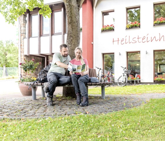 Deux personnes sont assises sur un banc devant la maison des pierres curatives à Einruhr, elles lisent une brochure. Des vélos sont garés devant le bâtiment, qui est décoré de fleurs., © -@Eifel Tourismus GmbH, AR-shapefruit-AG