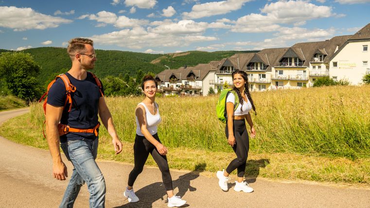 Eine Gruppe von drei Personen geht gemeinsam auf einemweg durch eine grüne Landschaft. Im Hintergrund sind moderne Gebäude unter einem blauen Himmel sichtbar.