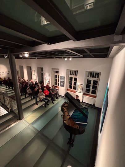 Ein Klavier steht auf einer Glasfläche, während eine Menge von Zuhörern auf Treppen sitzt. Die Atmosphäre ist konzertant und kreativ.