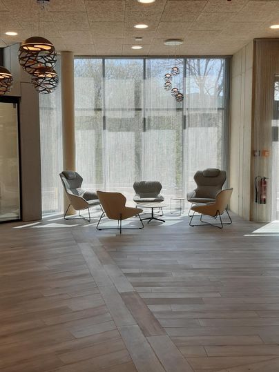 Ein moderner Innenraum mit Holzfußboden und großen Fenstern. In der Mitte stehen gemütliche Stühle und eine entspannte Atmosphäre herrscht.