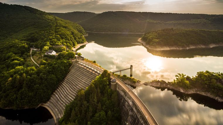Eine wunderschöne Landschaft mit einem Staudamm und einem ruhigen See. Umgeben von grünen Hügeln spiegelt sich das Licht der Sonne im Wasser.