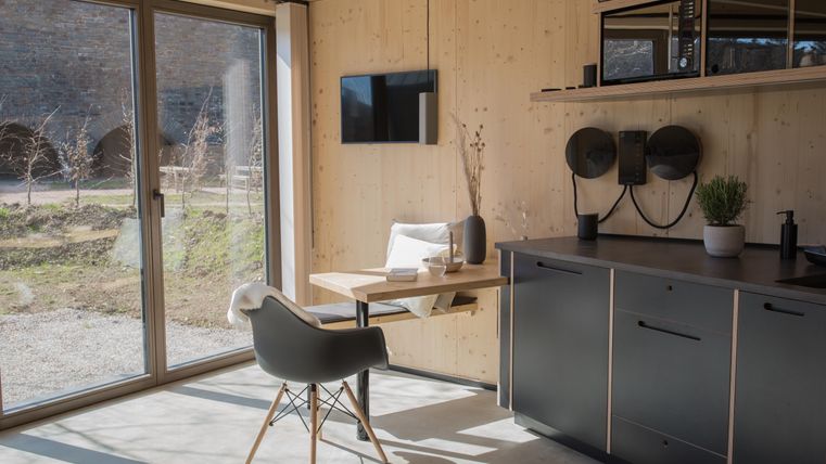 Ein modernes und helles Zimmer mit Holzwänden. Es gibt einen Tisch, einen Stuhl und eine Küchenzeile mit Geräten.