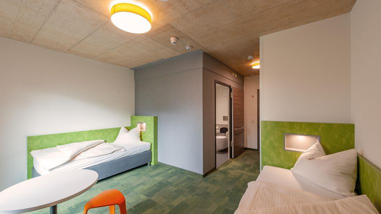 Ein modernes Zimmer mit zwei Betten und einem Tisch. Die Wände sind hell gestrichen und die Einrichtung hat farbenfrohe Akzenten.