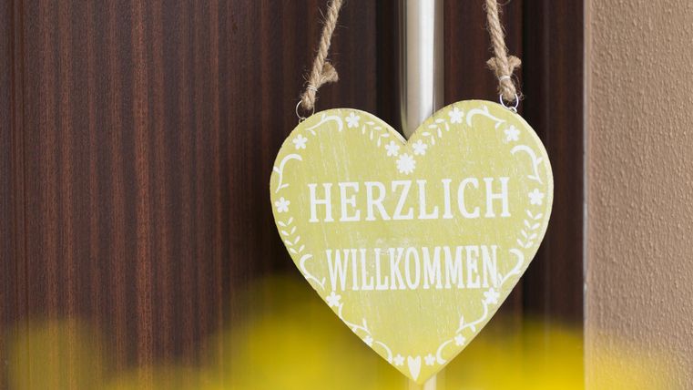 Ein herzförmiges Willkommensschild in sanften Gelbtönen. Darauf steht "Herzlich Willkommen" in weißer Schrift, aufgehängt an einer Tür.
