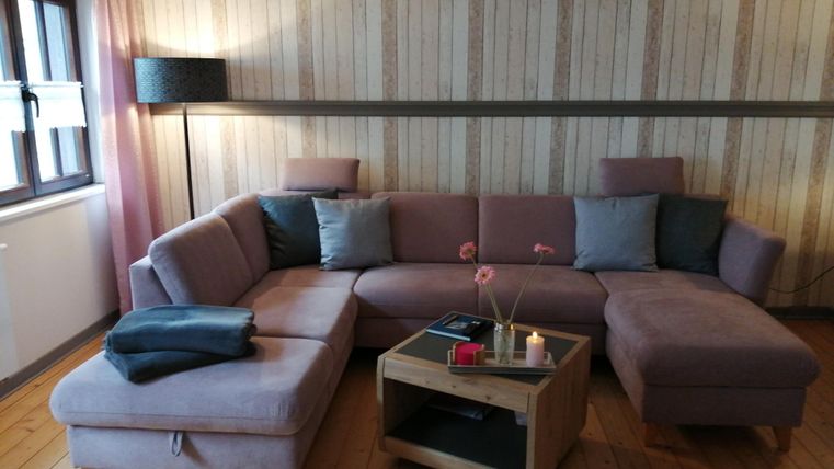 Ein gemütliches Wohnzimmer mit einer L-förmigen Couch in sanften Farben. Ein Holztisch und eine Stehlampe ergänzen die wohnliche Atmosphäre.