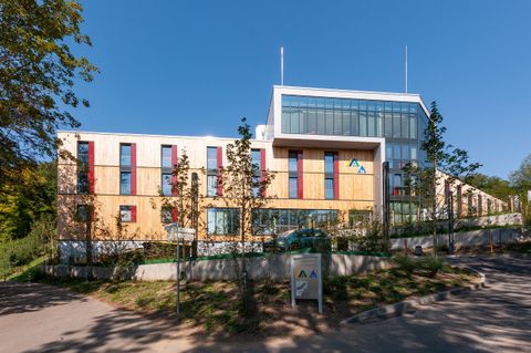 Ein modernes Gebäude mit großen Fenstern und bunten Fassaden. Die Umgebung ist grün und gut gestaltet.