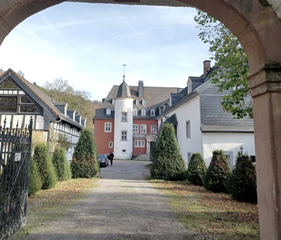 Blick durch ein Tor auf die Burg Dalbenden. Das Hauptgebäude ist von Bäumen umgeben, mit einem roten und weißen Turm in der Mitte., © Sweco GmbH