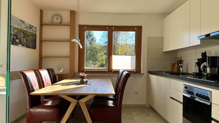 Eine moderne Küche mit einem Essbereich. Der Tisch aus Holz steht zwischen vier roten Stühlen und großen Fenstern, die viel Tageslicht hereinlassen.