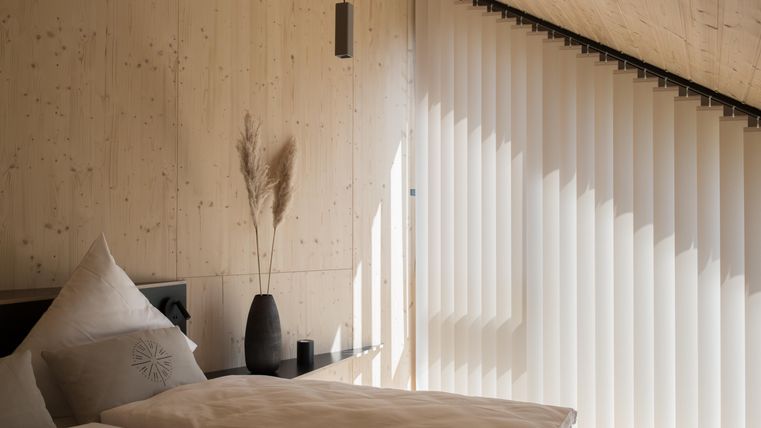 Ein modernes Schlafzimmer mit hellen Wänden aus Holz und einem großen Fenster. Sanftes Licht fällt durch die Jalousien auf das Bett und die Dekoration.