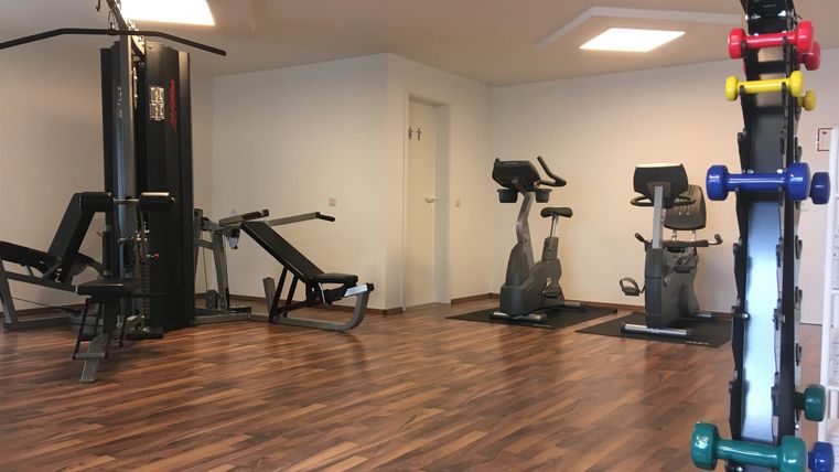 Ein modernes Fitnessstudio mit verschiedenen Trainingsgeräten. Im Vordergrund stehen Hanteln und im Hintergrund sind Ergometer und eine Kraftstation zu sehen.