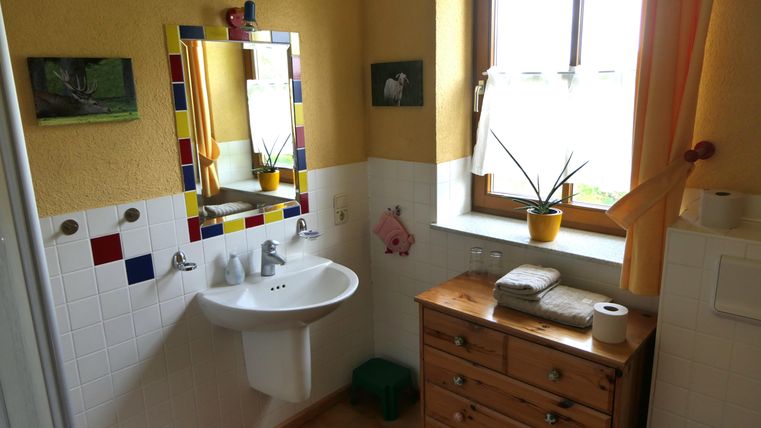 Ein helles Badezimmer mit gelben Wänden und einem großen Fenster. Es gibt ein Waschbecken, einen Holztisch mit Handtüchern und eine Pflanze auf der Fensterbank.