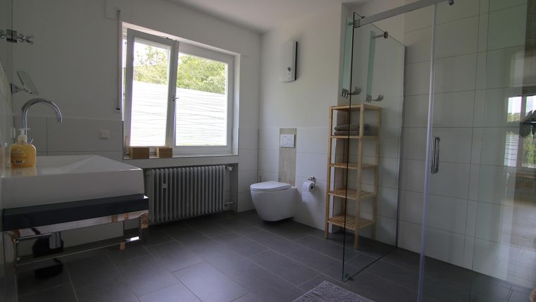 Ein modernes Badezimmer mit einer großen Glasdusche und einem eleganten Waschbecken. Es gibt ein Fenster, das viel Licht hineinlässt, sowie einen Holzregal für Handtücher.