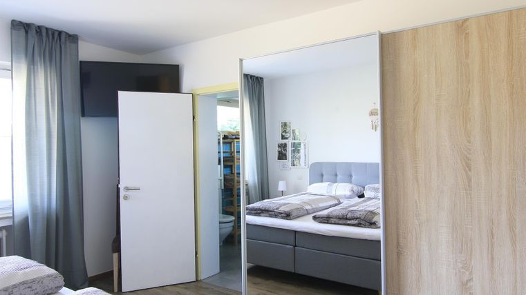 Ein helles Schlafzimmer mit einem Doppelbett und grau gestreiften Bettbezügen. Neben dem Bett steht ein Spiegel und es gibt Vorhänge vor dem Fenster.