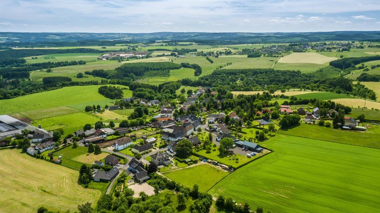 Eine Luftaufnahme eines malerischen Dorfes umgeben von grünen Feldern und Hügeln. Der Himmel ist klar mit wenigen Wolken.