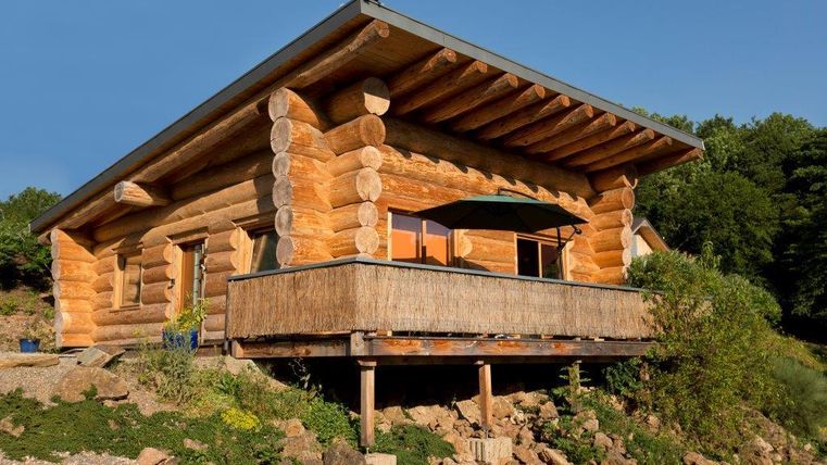 Ein gemütliches Blockhaus mit einer großen Veranda. Umgeben von grüner Natur und klarem Himmel.