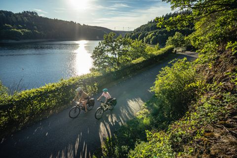 Zwei Radfahrer fahren auf einem Weg entlang eines Sees bei Sonnenschein, umgeben von grüner Natur.